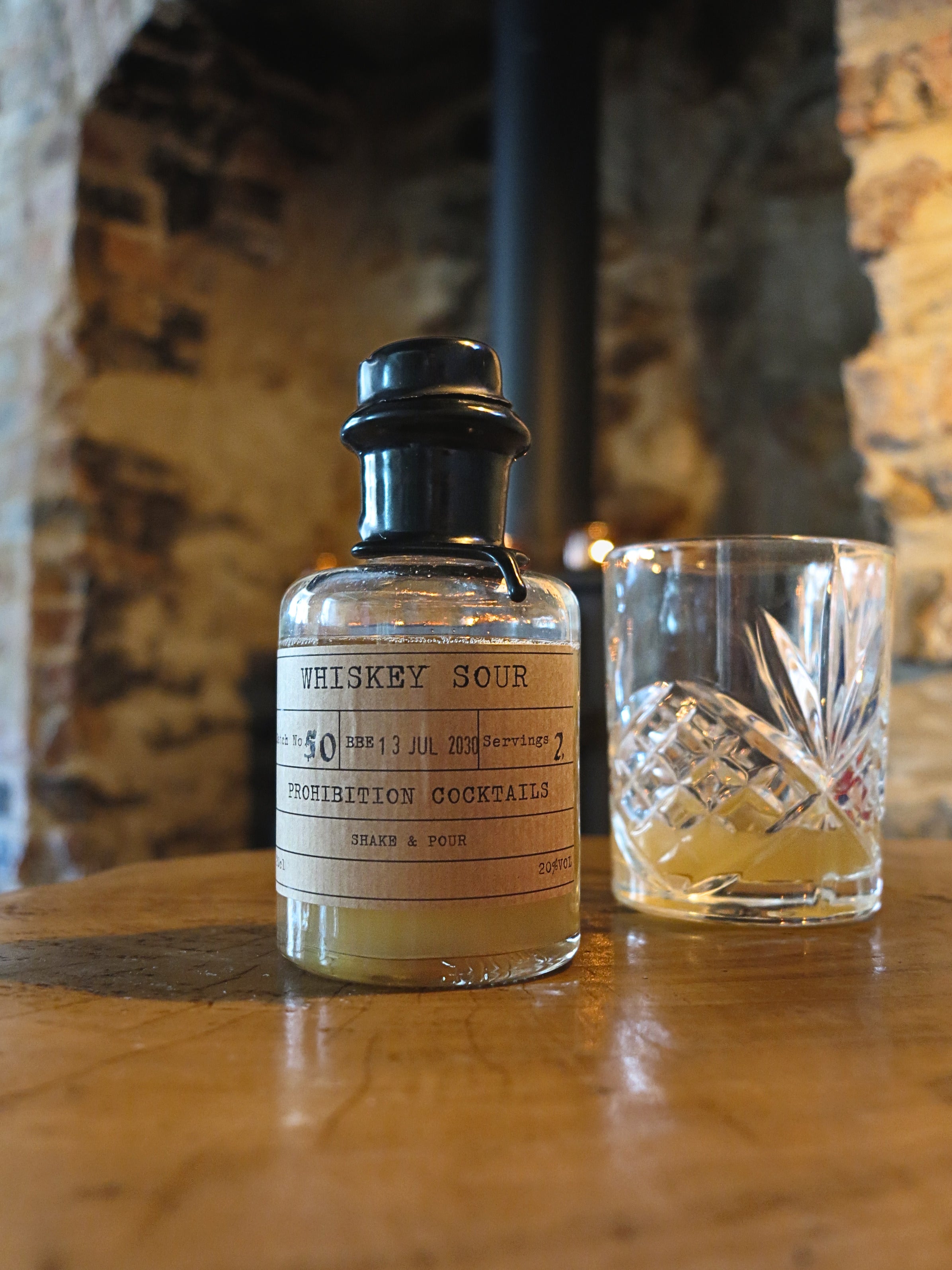 A bottle of Whiskey Sour cocktail mix with a black cap and cream-colored label, placed on a wooden table next to a clear cut-glass rocks tumbler containing a golden liquid, with a rustic stone archway in the background.