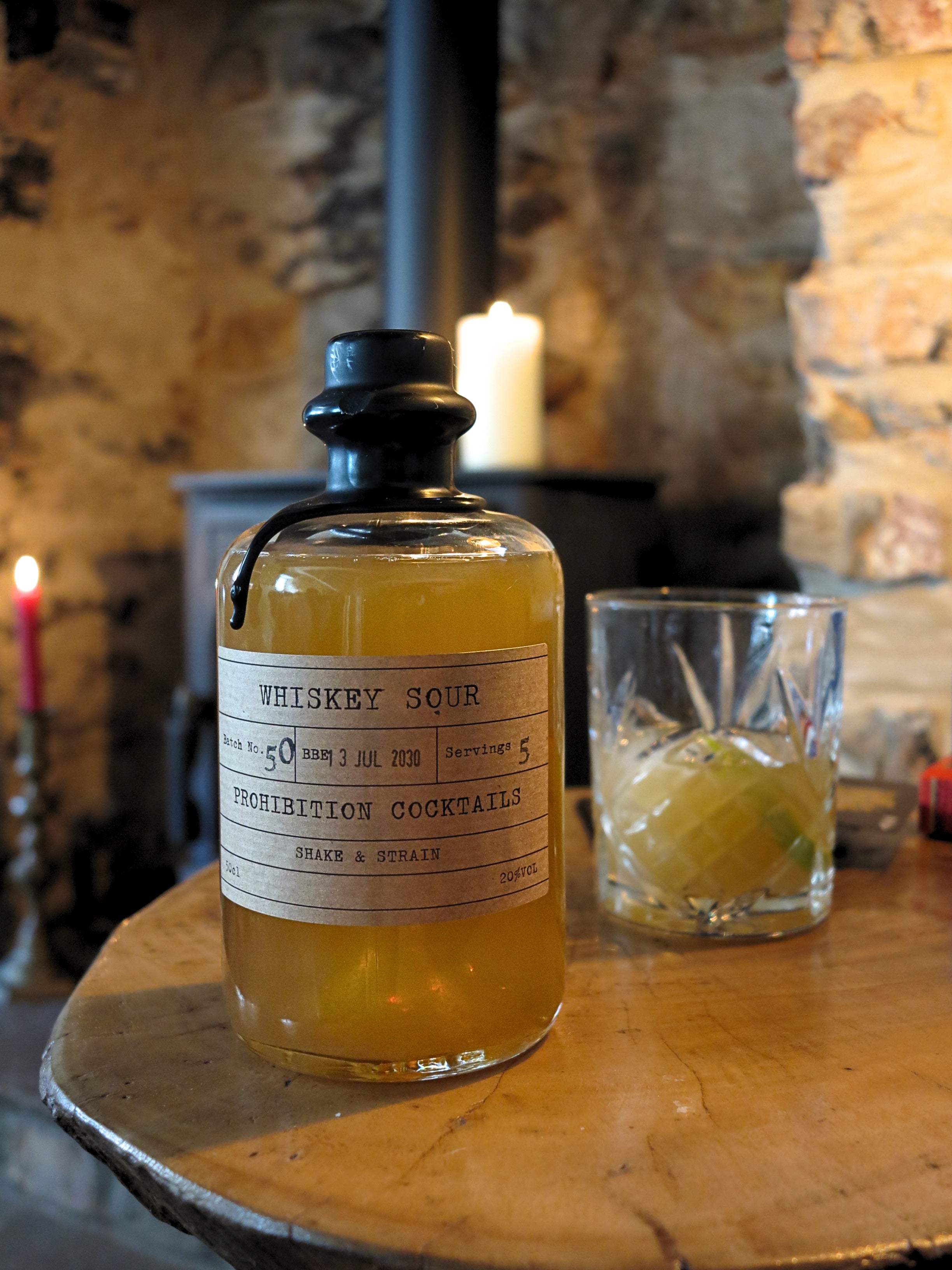 A bottle of Prohibition Cocktails Whiskey Sour with a cream label and black cap, displayed on a rustic wooden table next to a glass of the drink in a cozy, dimly lit stone fireplace setting.